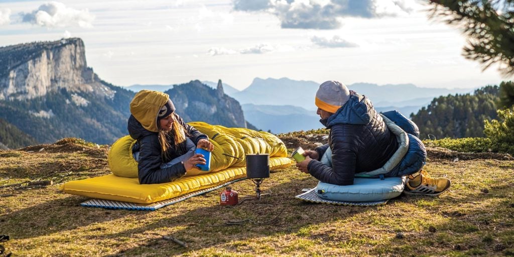 Two people laying in Therm-A-Rest sleeping bags and pads sharing a meal.